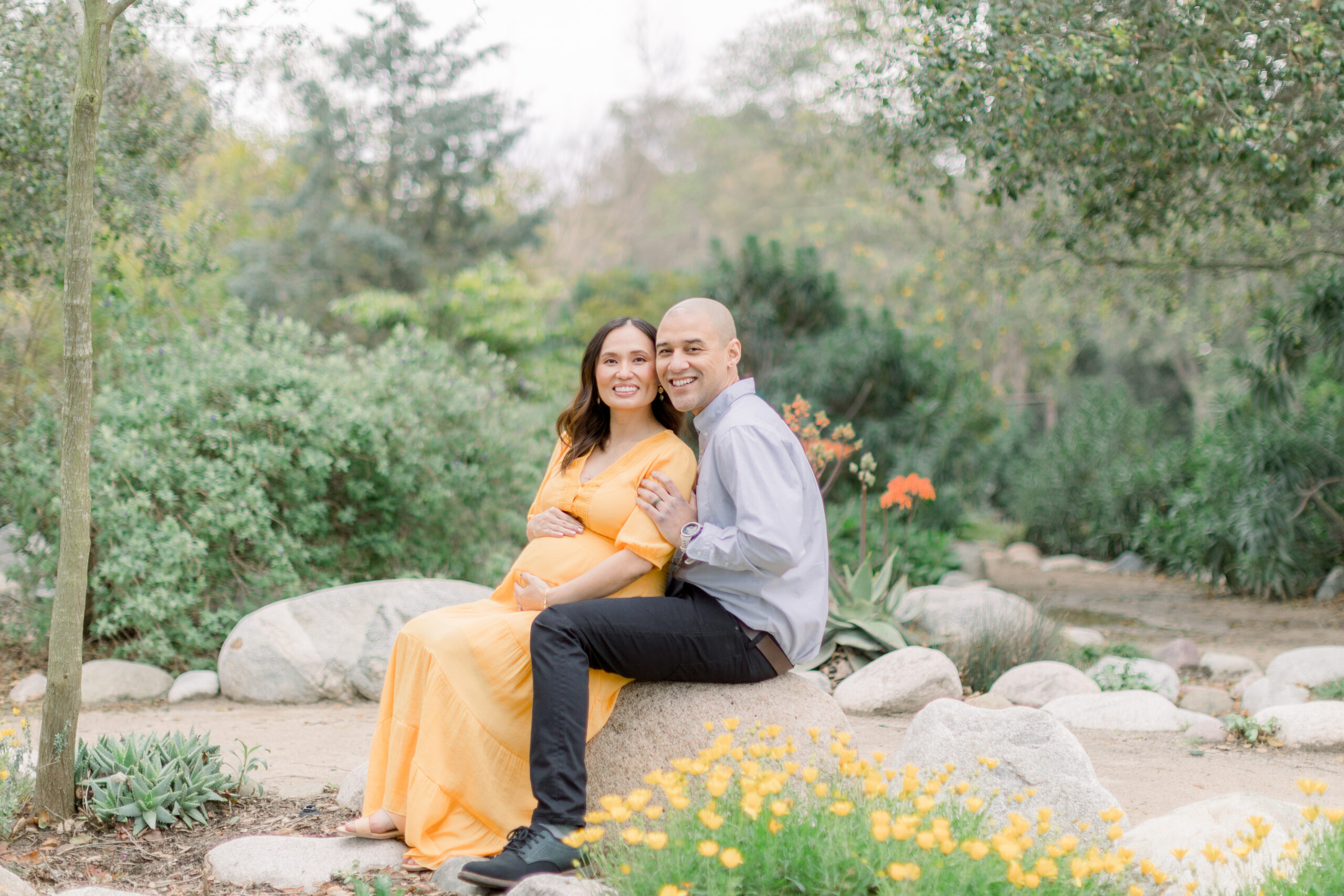 Smiling expecting couple sitting on a rock surrounded by greenery and wildflowers during a maternity session at the Secret Garden in Huntington Central Park, Huntington Beach