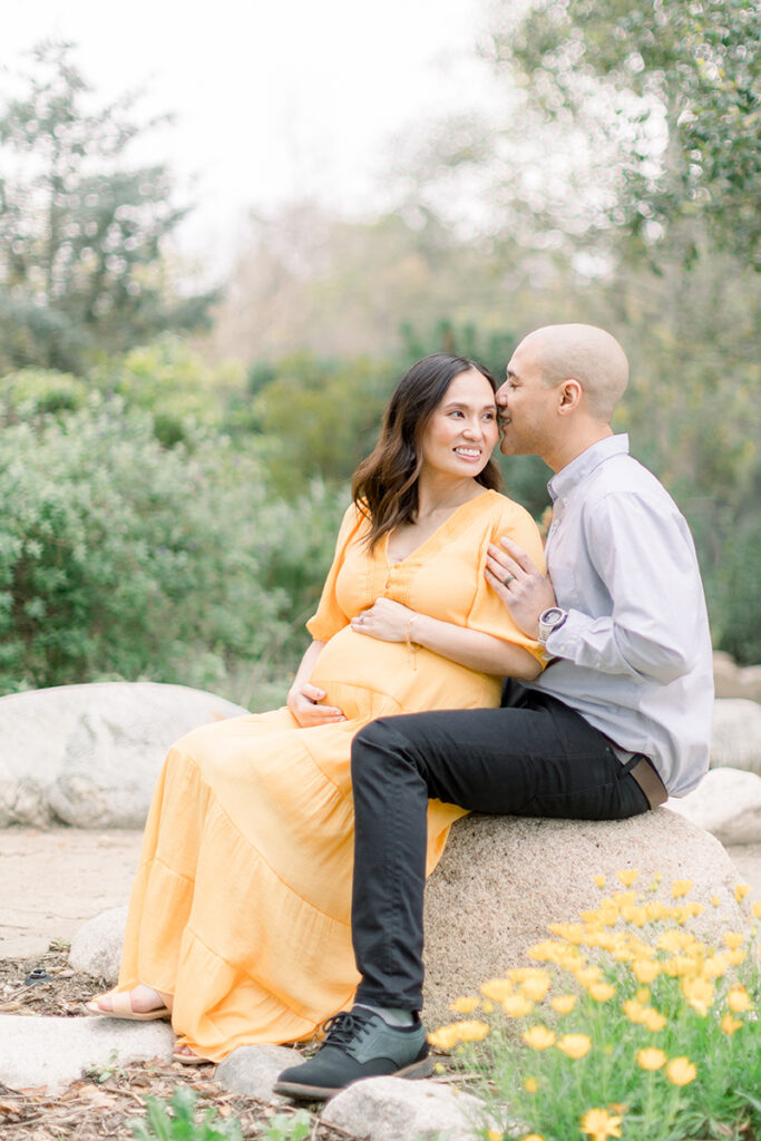 Husband kissing his pregnant wife on the temple during a maternity session at the Secret Garden in Huntington Central Park, with wildflowers blooming around them
