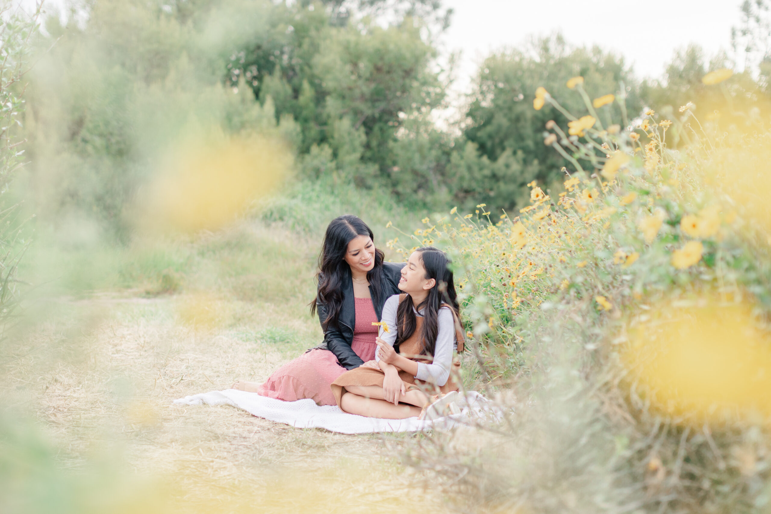 Mother and daughter sitting on a blanket in a San Diego wildflower field, smiling and enjoying a sweet moment together during a spring mini session.