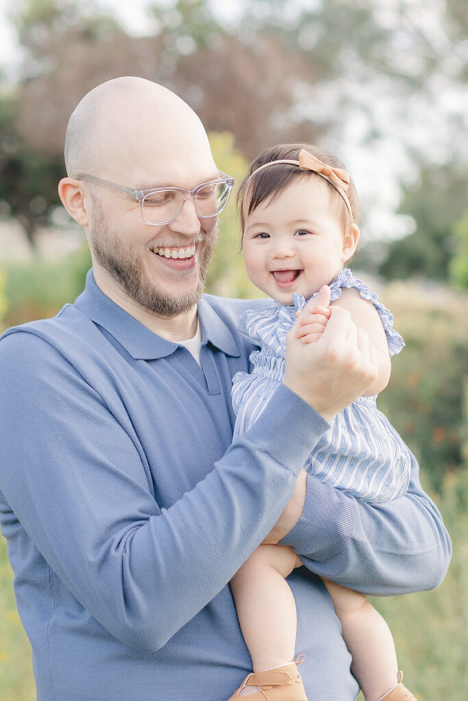 Father laughing while holding his smiling baby girl during a spring family photo session in San Diego, both wearing soft blue tones with greenery in the background