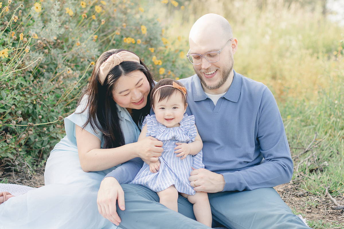 Happy family of three sitting together in a field of wildflowers during a spring photo session in San Diego, smiling and enjoying a tender moment in soft natural light