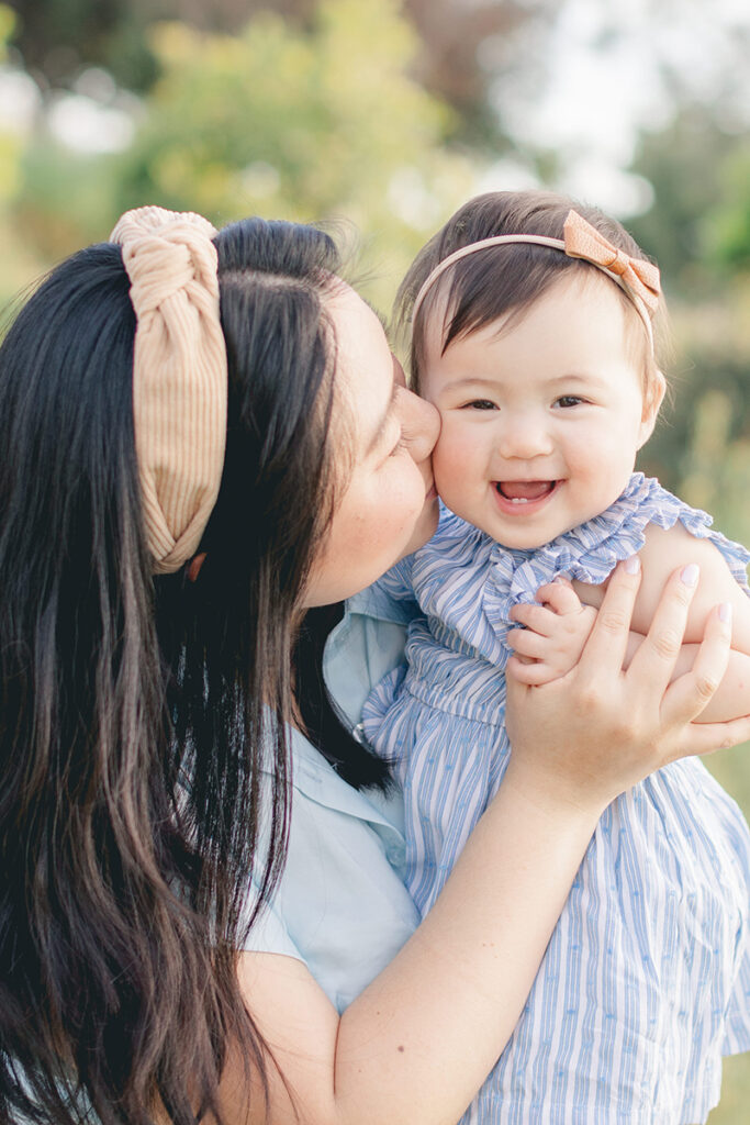 Sweet moment of a mother kissing her smiling baby girl on the cheek during a spring photo session in San Diego, surrounded by soft light and natural greenery