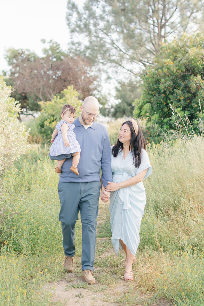 Family walking through a sunlit park trail during a spring photo session in San Diego, dressed in soft blue tones with natural light and greenery all around