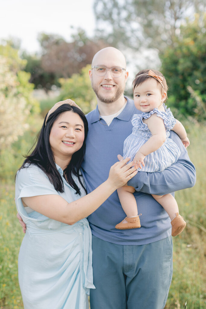 Happy family of three during a spring mini session in San Diego, smiling at the camera while surrounded by soft greenery and natural light