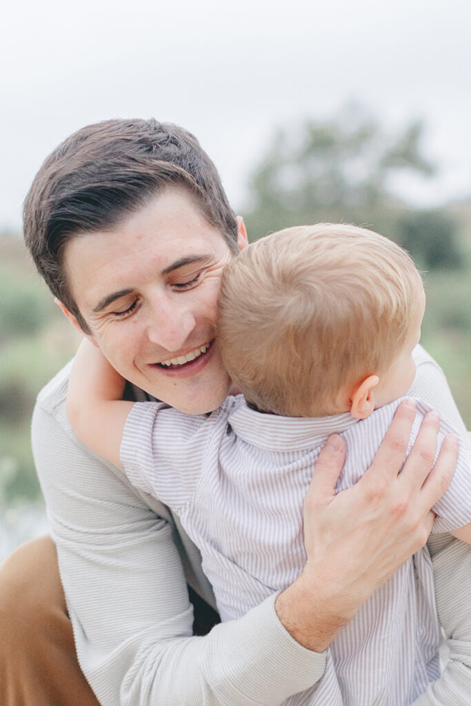 Dad smiling with eyes closed while hugging his toddler son during an emotional moment at Kit Carson Park in San Diego