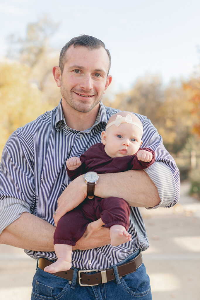 Father holding his baby girl dressed in maroon with a cream bow headband, smiling during a sunny fall family session in Santee, California.