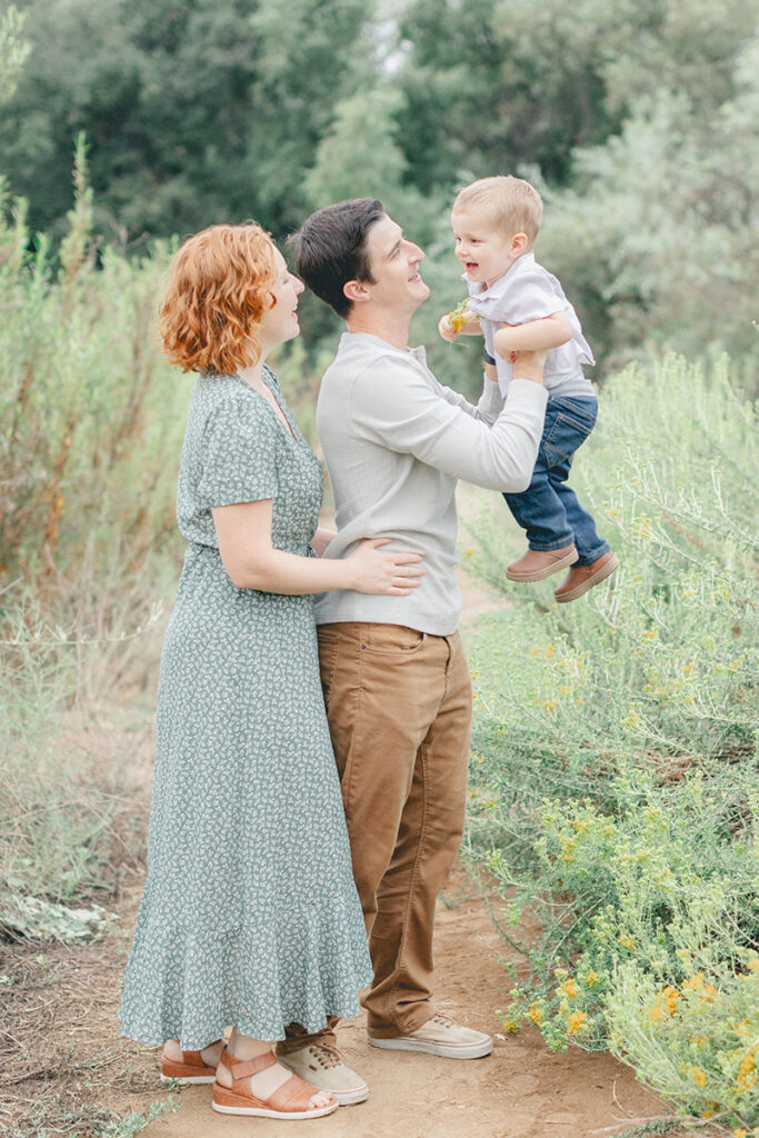 Dad lifting toddler son into the air while mom smiles during a joyful family session at Kit Carson Park in San Diego
