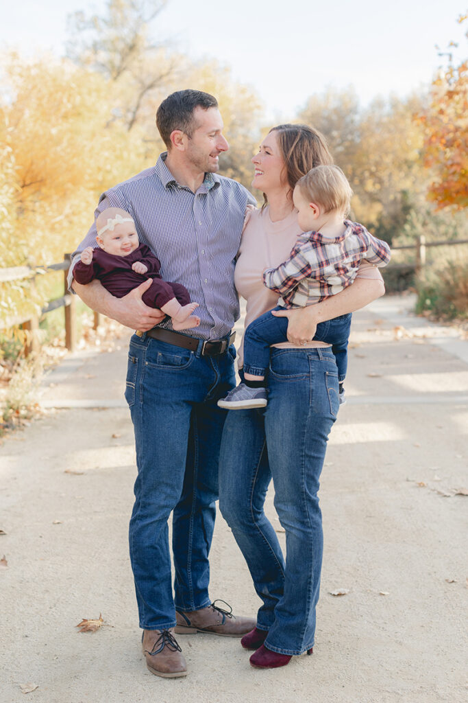 Smiling family of four standing together on a sunlit path in Santee, California, surrounded by golden fall foliage during a candid autumn photo session.