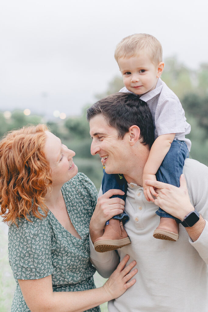 Family smiling together as dad carries toddler son on his shoulders during a sunset photo session at Kit Carson Park in San Diego