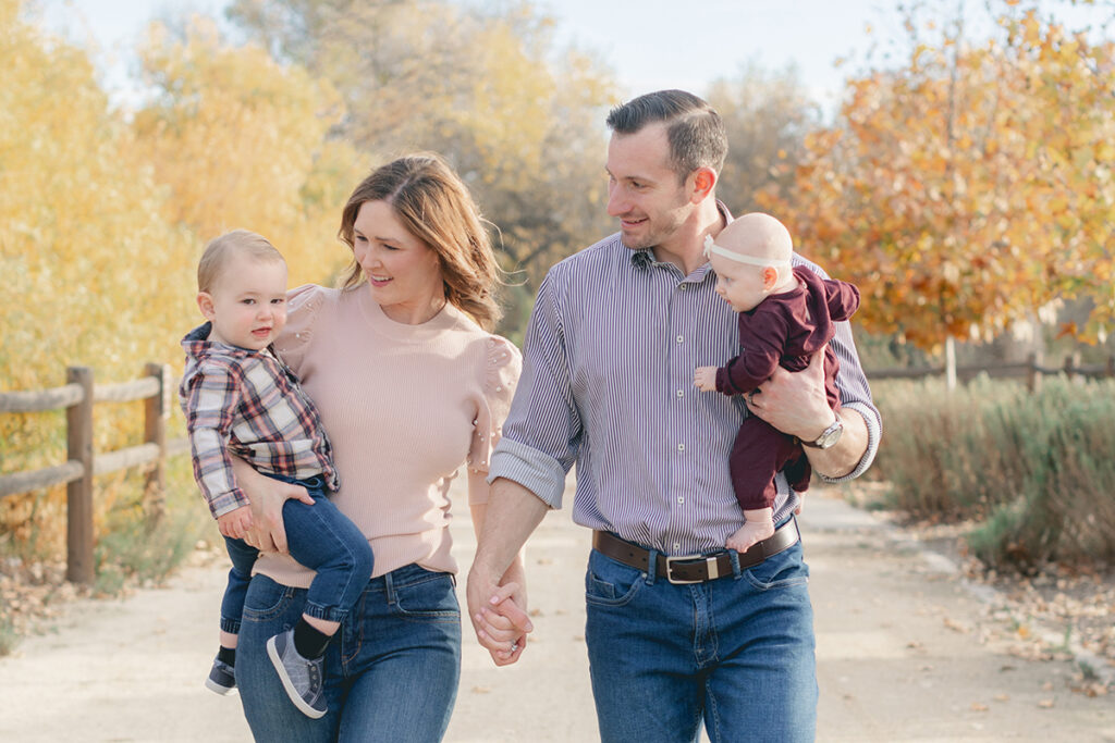 Family of four walking hand in hand along a sunlit trail in Santee, California, surrounded by golden fall foliage during a light-filled mini session.