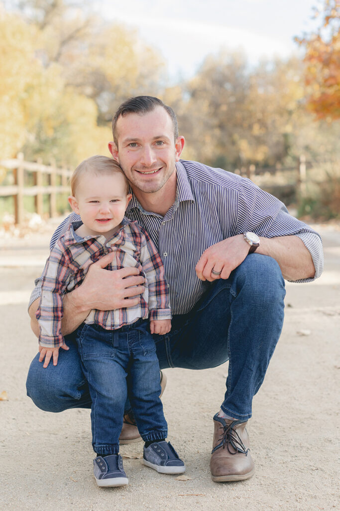 Father and toddler son smiling together on a tree-lined path during a fall family photo session in Santee, California, with golden foliage in the background.