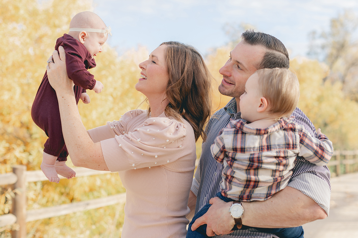 Smiling mom lifting baby girl into the air while dad holds toddler, captured during a fall family photo session in Santee.