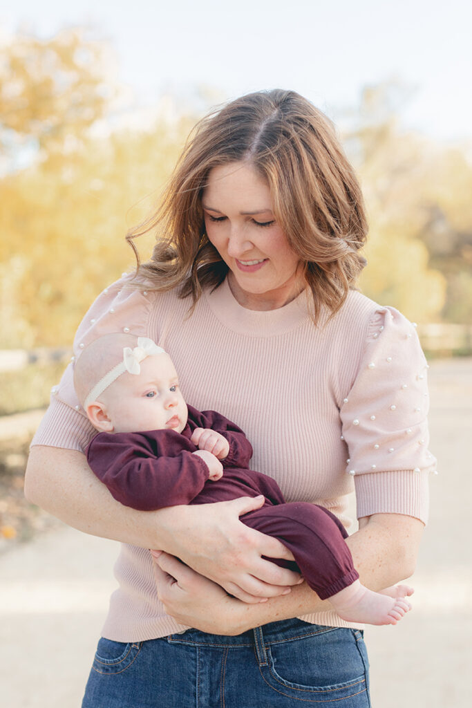 Mother gently holding her baby girl dressed in maroon, both bathed in soft natural light during a fall family photo session in Santee, California.