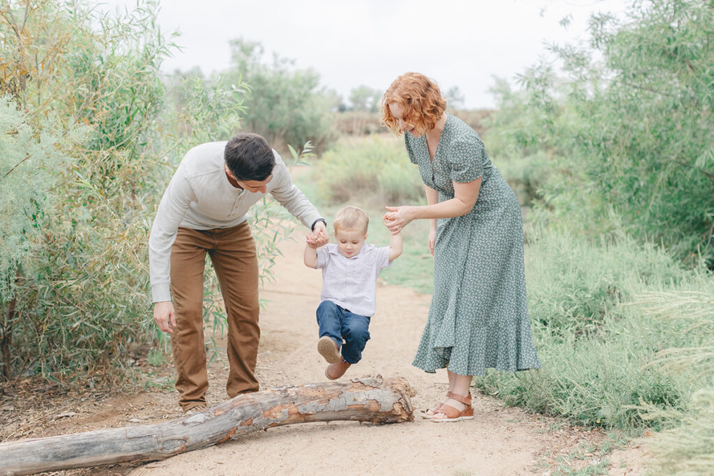 Parents helping their toddler jump over a log during a playful family photo session at Kit Carson Park in San Diego