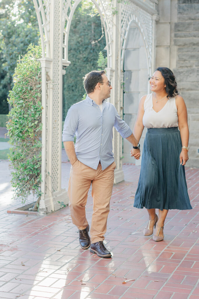 Cute couple walking hand in hand under an arched trellis at Greystone Mansion during their engagement session in Beverly Hills, smiling at each other with joy.
