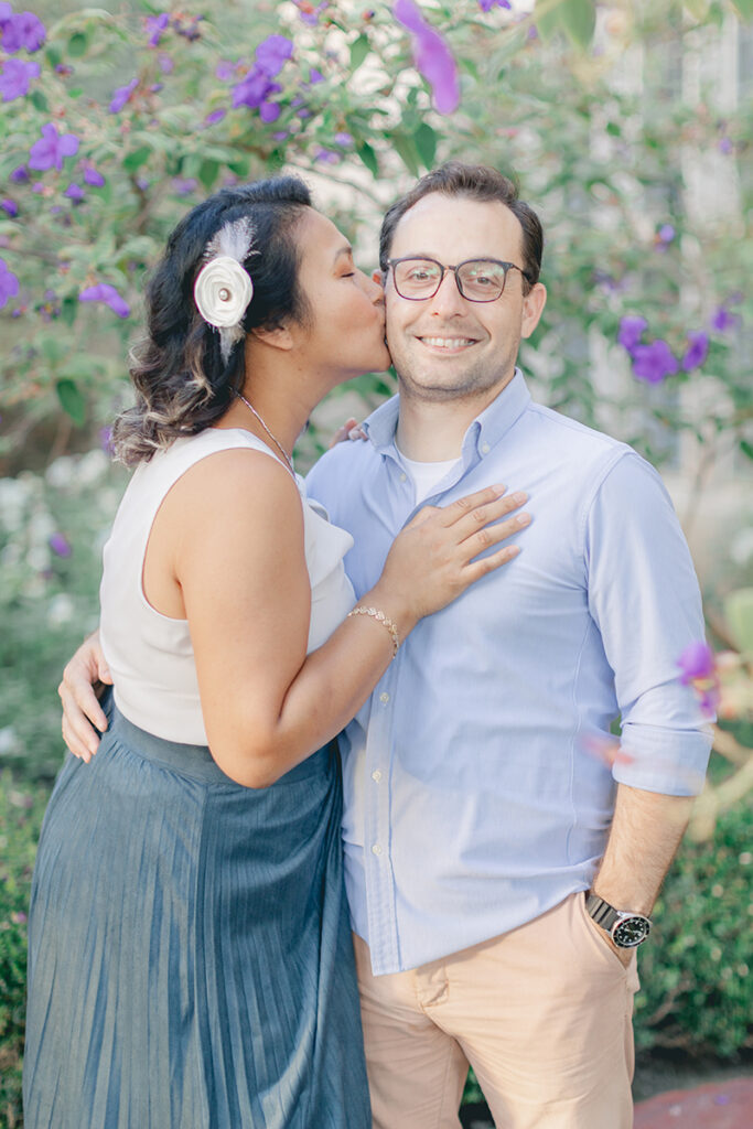Woman kissing her fiancé on the cheek while he smiles softly, surrounded by blooming purple flowers during their engagement session at Greystone Mansion in Beverly Hills.