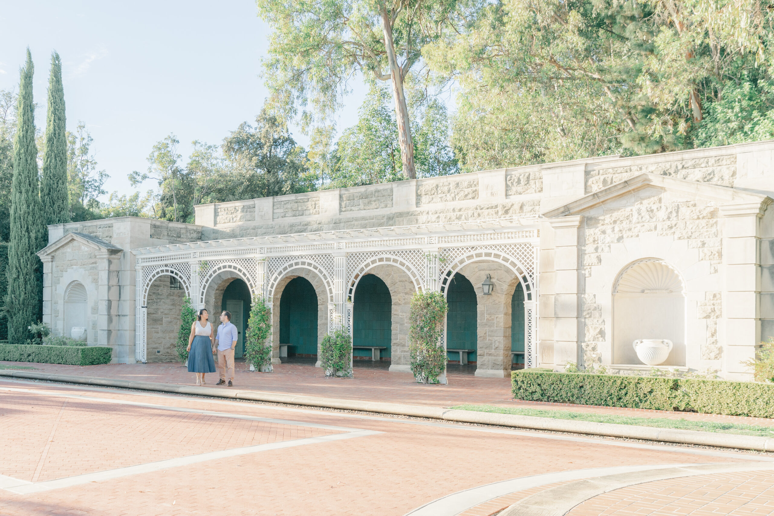 Engaged couple standing hand in hand in front of the grand stone arches and garden architecture at Greystone Mansion in Beverly Hills during their romantic engagement session.