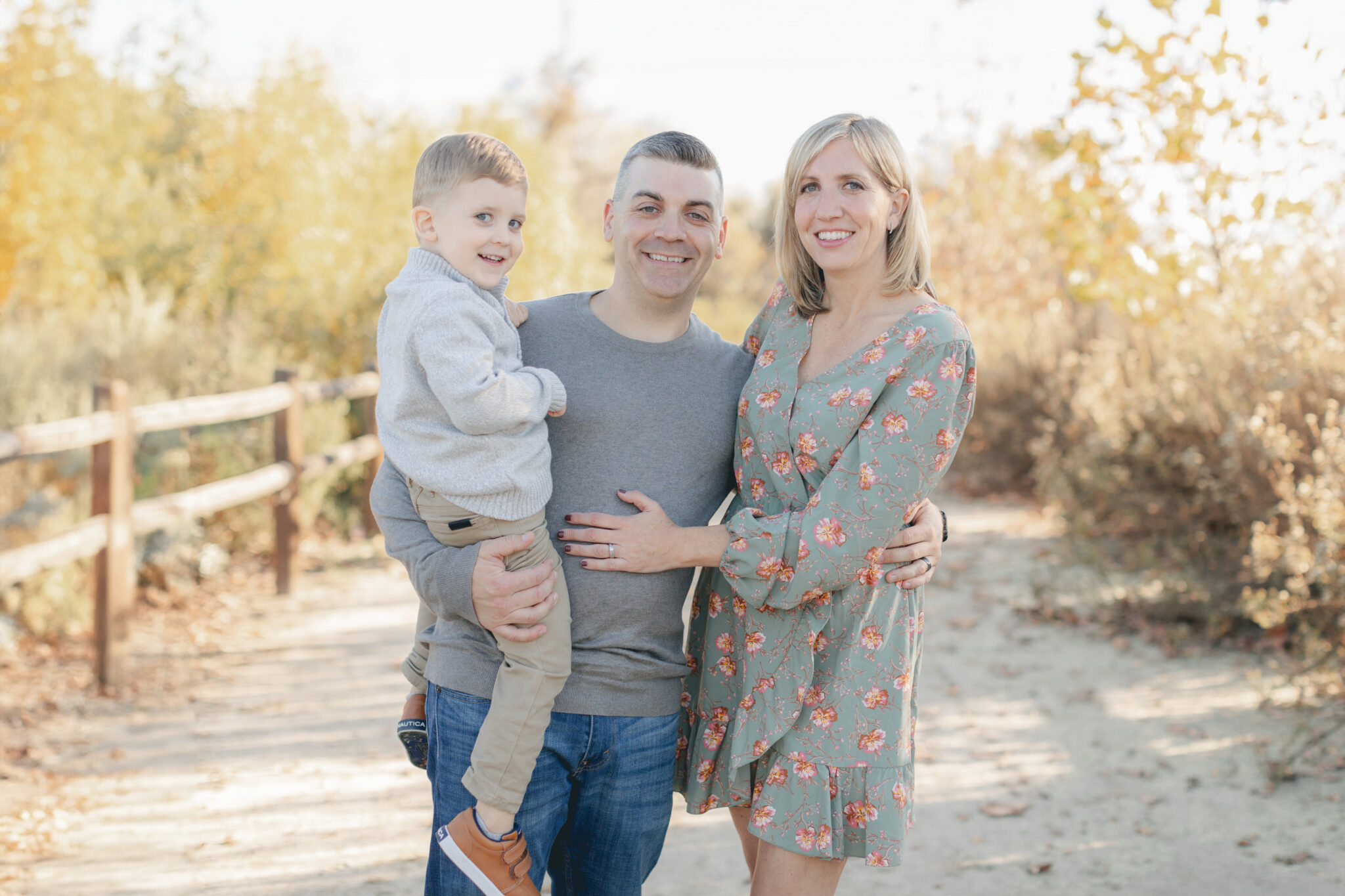 Smiling family of three during a fall mini session in San Diego, standing on a sunlit nature trail with autumn leaves