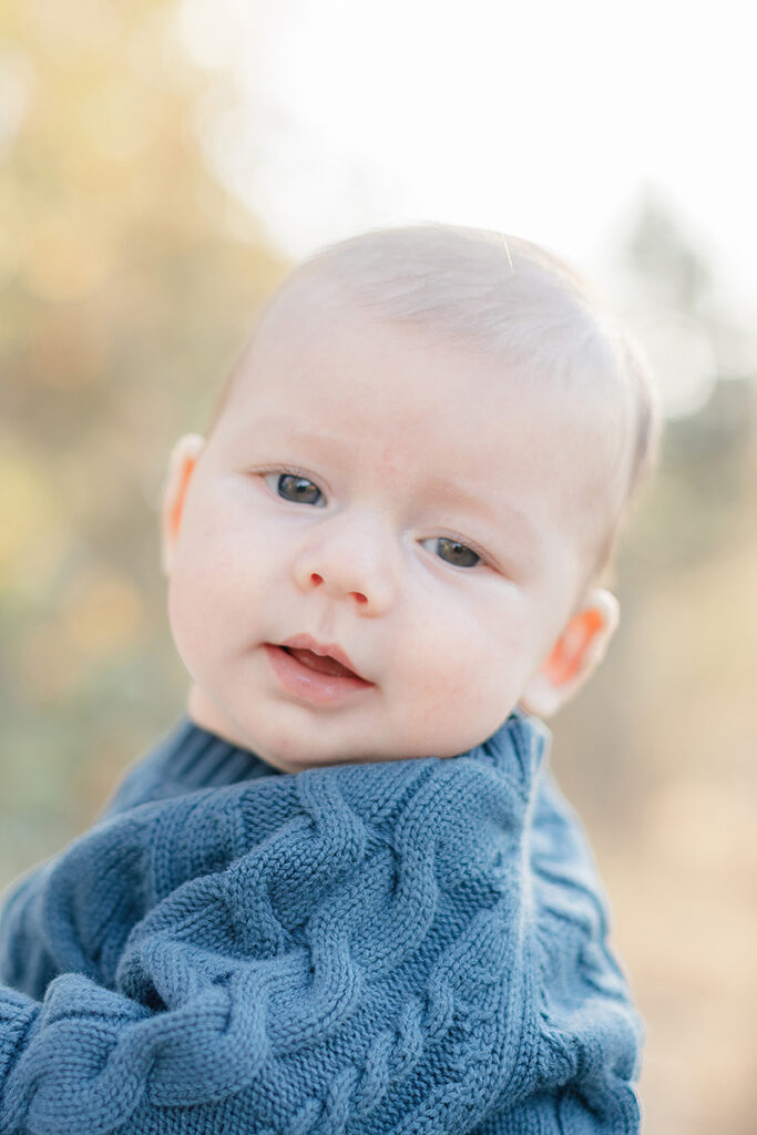 Close-up portrait of a 3-month-old baby in a blue knit sweater during a golden hour photo session in La Mesa, San Diego, with soft natural light and blurred autumn background.