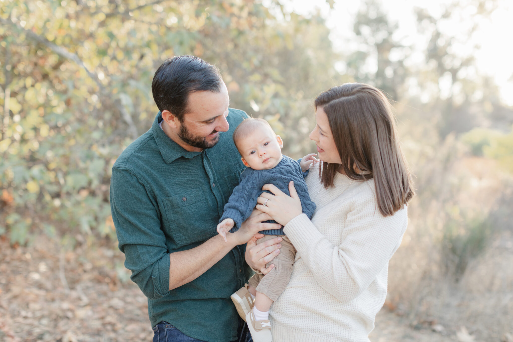 Parents smiling at their baby during a golden hour family session in La Mesa, San Diego, surrounded by soft sunlight and autumn foliage.