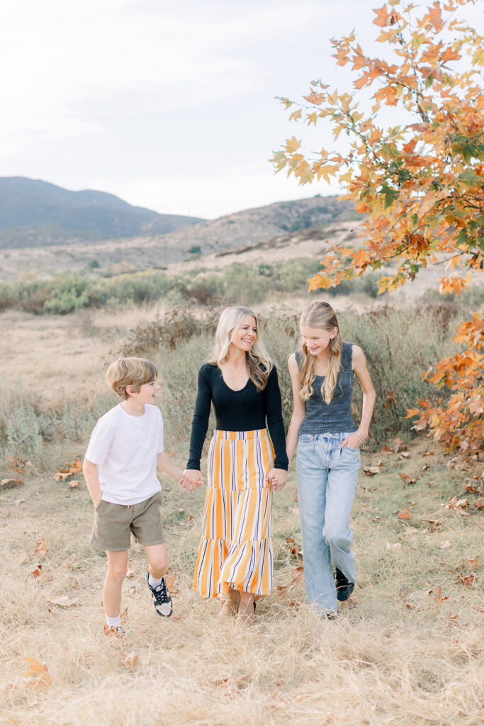 Mom walking hand-in-hand with her two children during a candid fall family photo session in San Diego with mountain views and autumn leaves