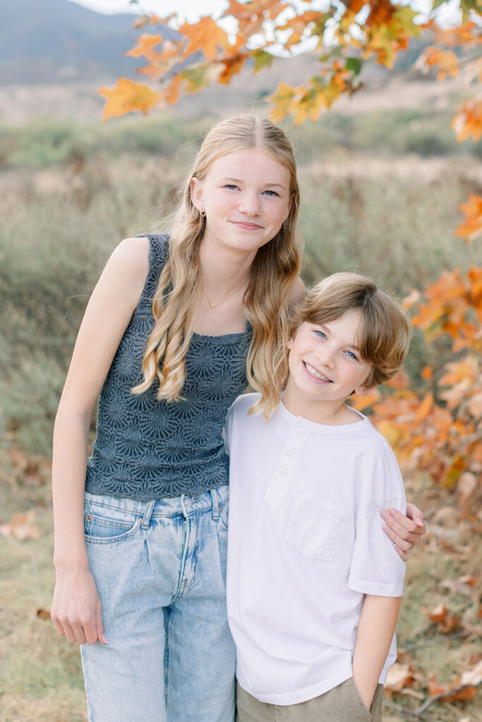 Brother and sister smiling and posing together during a fall family photo session in San Diego with autumn leaves in the background