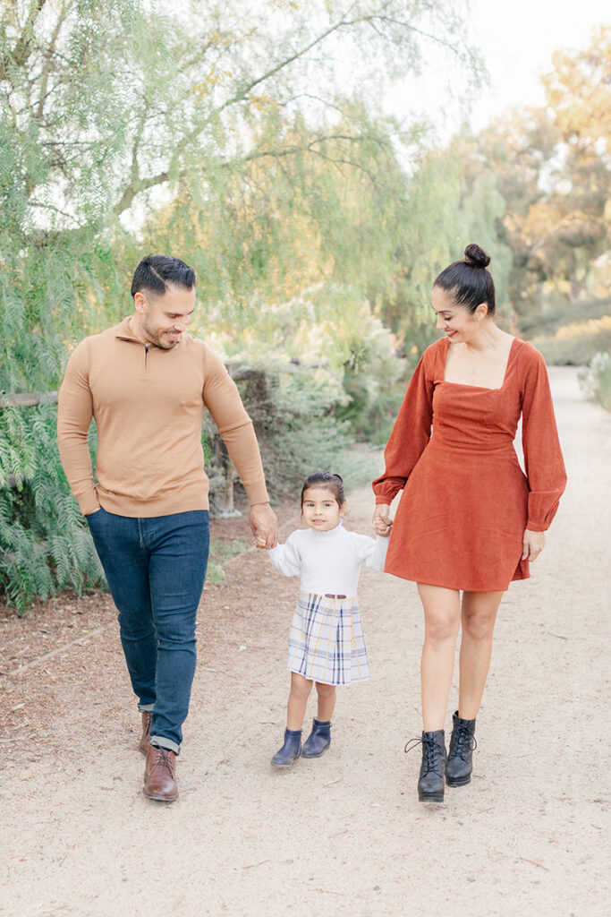 Family of three walking hand in hand along a nature trail at Montevalle Park in Chula Vista, dressed in warm fall colors during an outdoor photoshoot