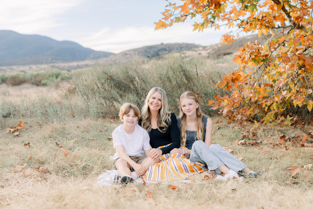 Smiling mom with her two kids sitting on a blanket under a tree with fall leaves during a family photo session in San Diego