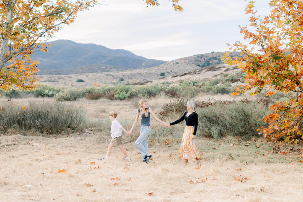 Mom and her two kids holding hands and walking through an open field with mountain views and fall foliage during a family photo session in San Diego