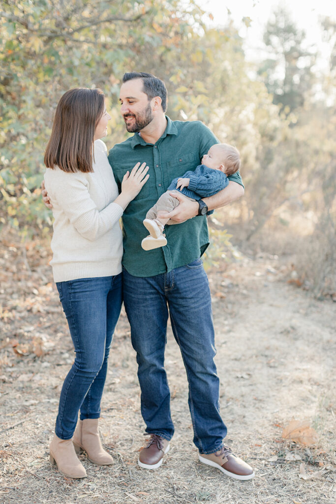 Happy parents standing close together while dad holds their baby during a golden hour family session in La Mesa, San Diego, surrounded by fall foliage and soft sunlight.