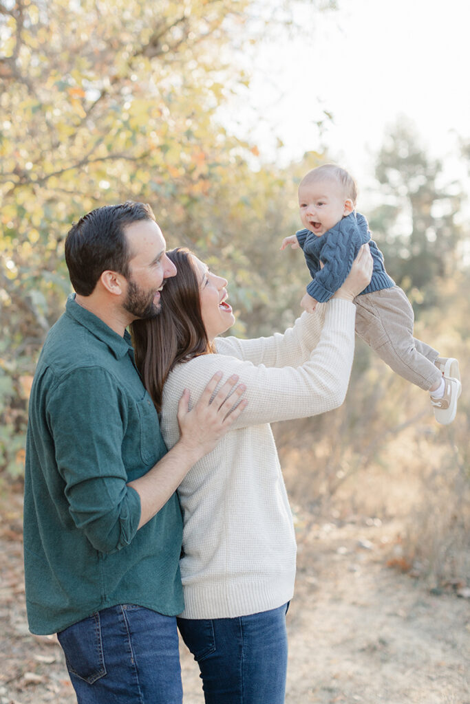 Joyful candid moment of parents smiling as mom lifts their baby into the air during golden hour at Harry Griffen Park in La Mesa, San Diego, with warm sunlight and fall colors in the background.