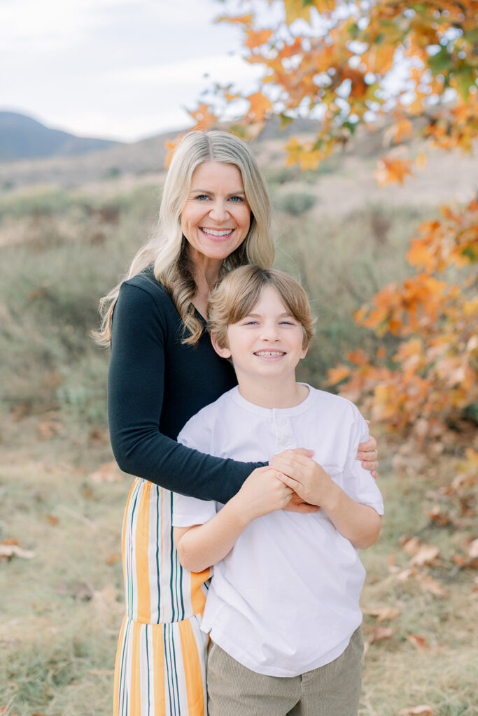 Smiling mom holding her son during a fall photo session in San Diego with autumn trees and mountain views in the background