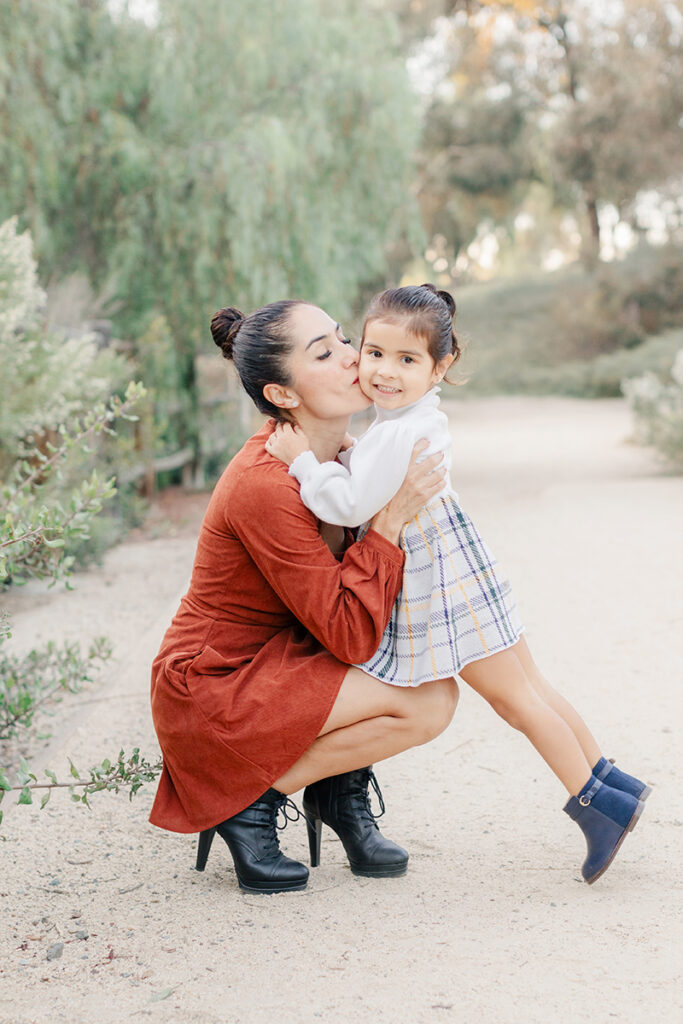 Mother kneeling down to kiss her smiling daughter on the cheek during a family photoshoot at Montevalle Park in Chula Vista