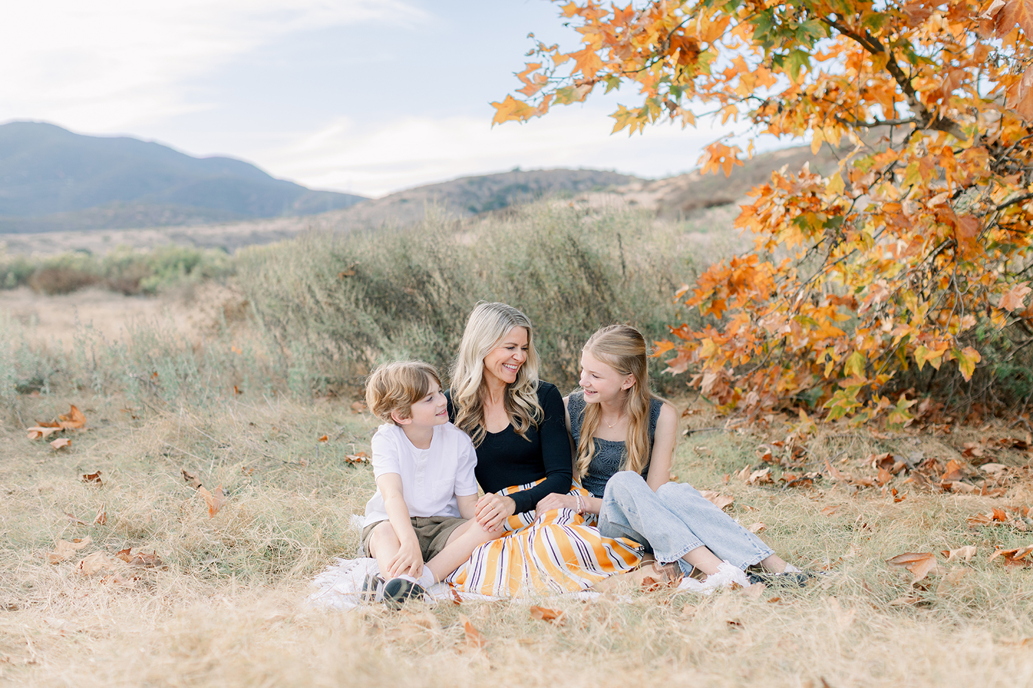 Mom with two children sitting under a tree with fall leaves during a natural light family photo session in San Diego