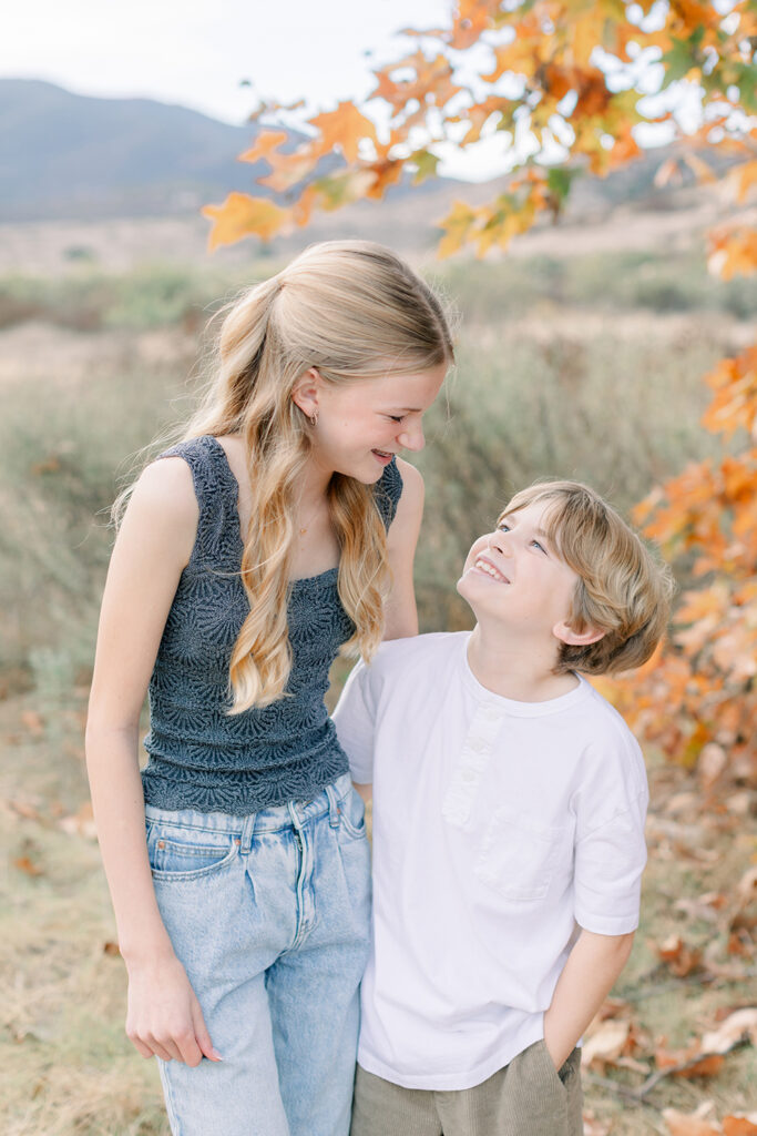 Brother and sister smiling at each other during a fall photo session in San Diego with autumn leaves and soft mountain views in the background