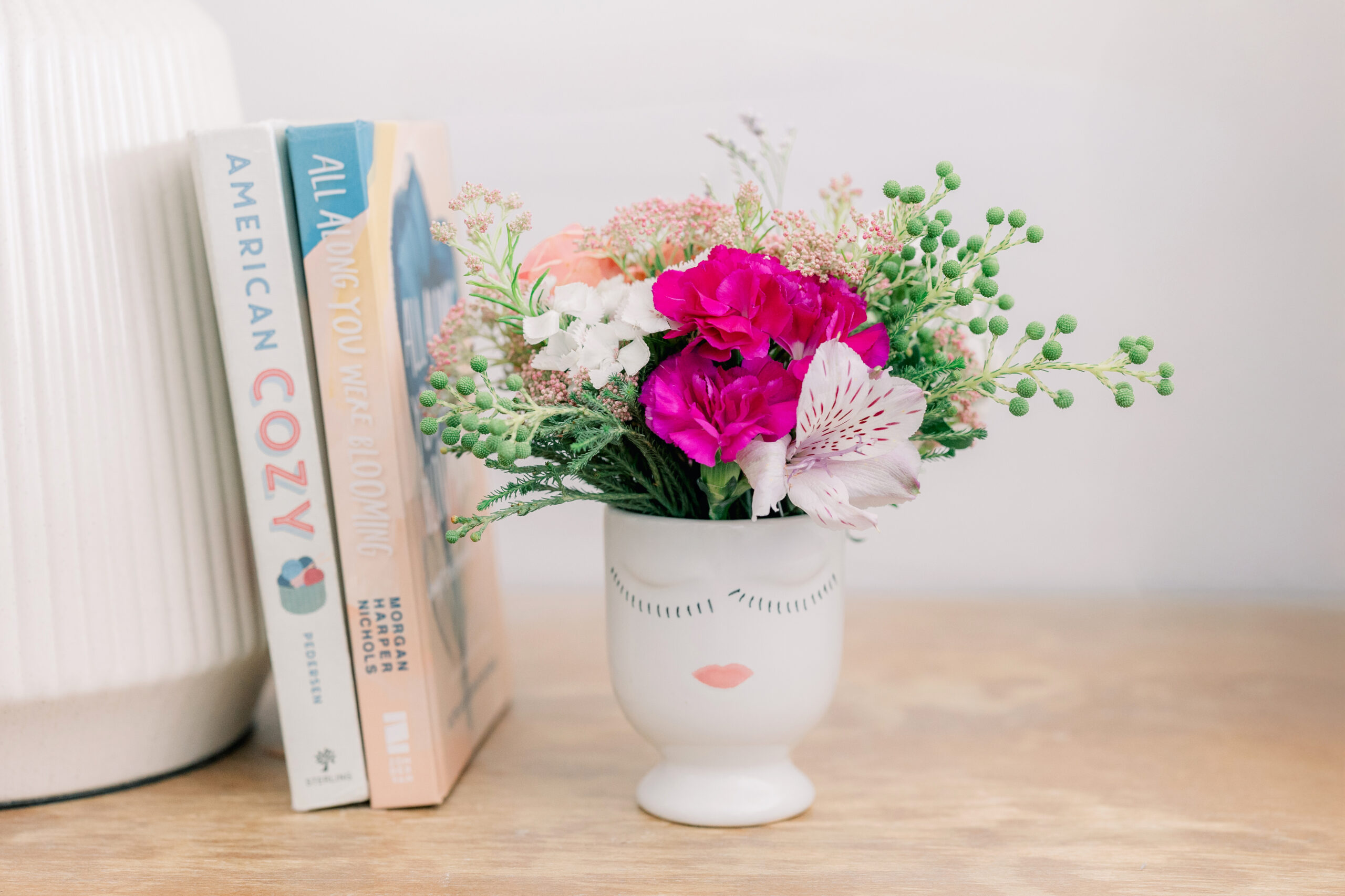 Bright floral arrangement in a minimalist face vase on a wooden table with books titled "American Cozy" and "All Along You Were Blooming" in the background, styled in a cozy, modern home setting.
